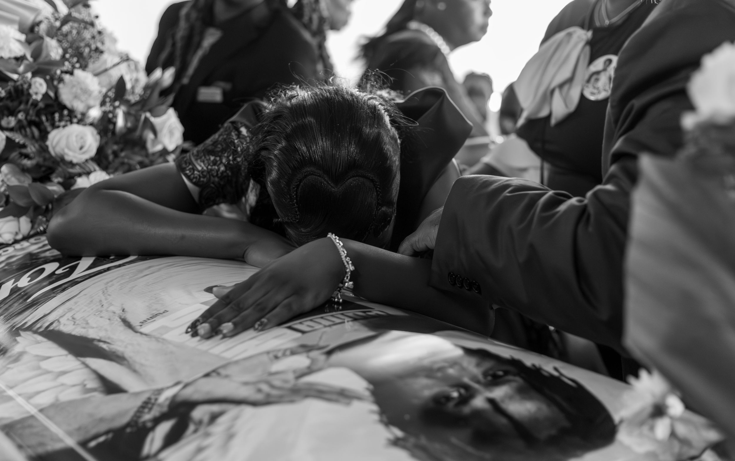 Black and white photo of a grieving woman leaning over a casket at a funeral service.