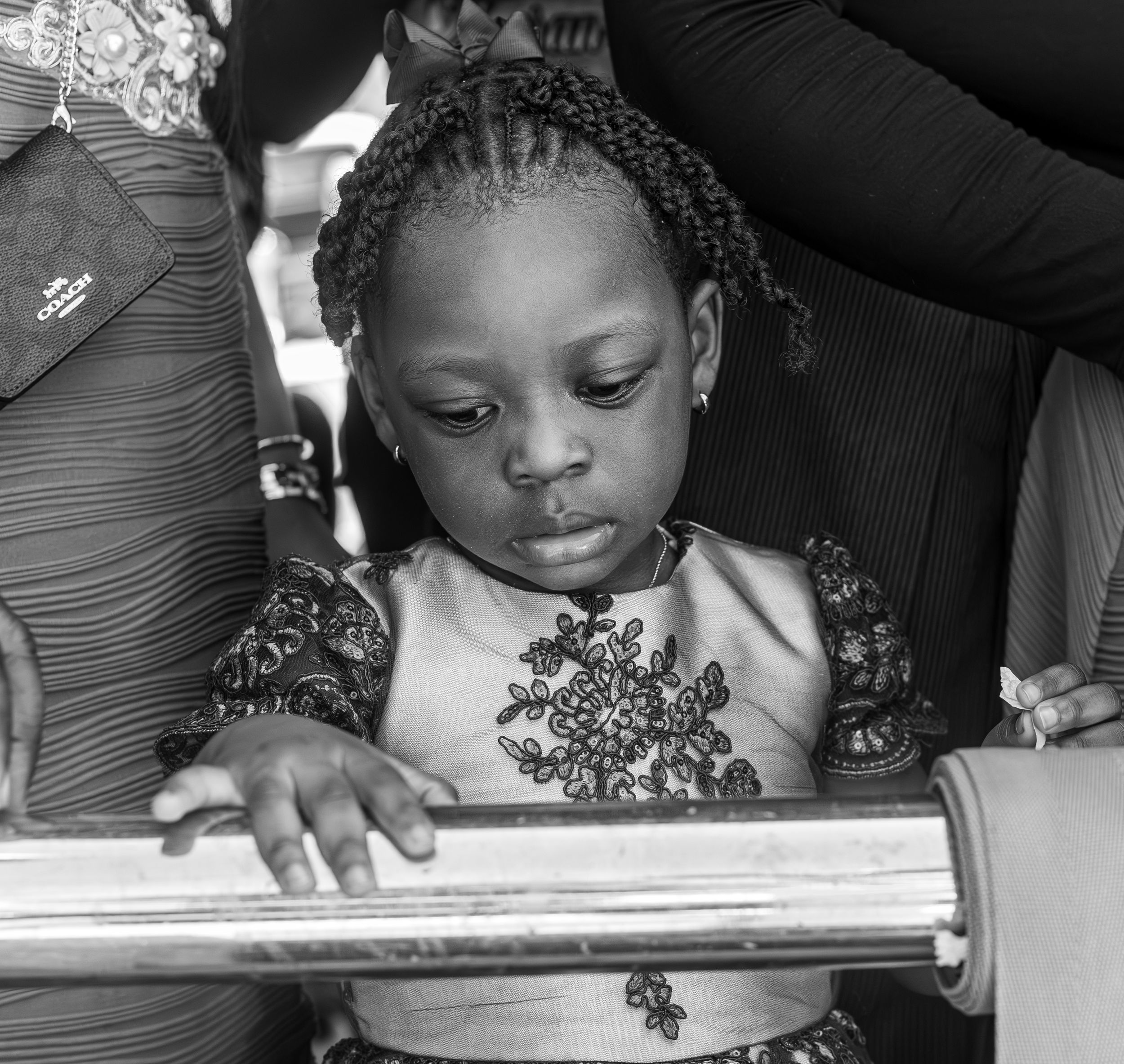 Black and white portrait of a young African girl with braids wearing an embroidered dress, looking down thoughtfully.