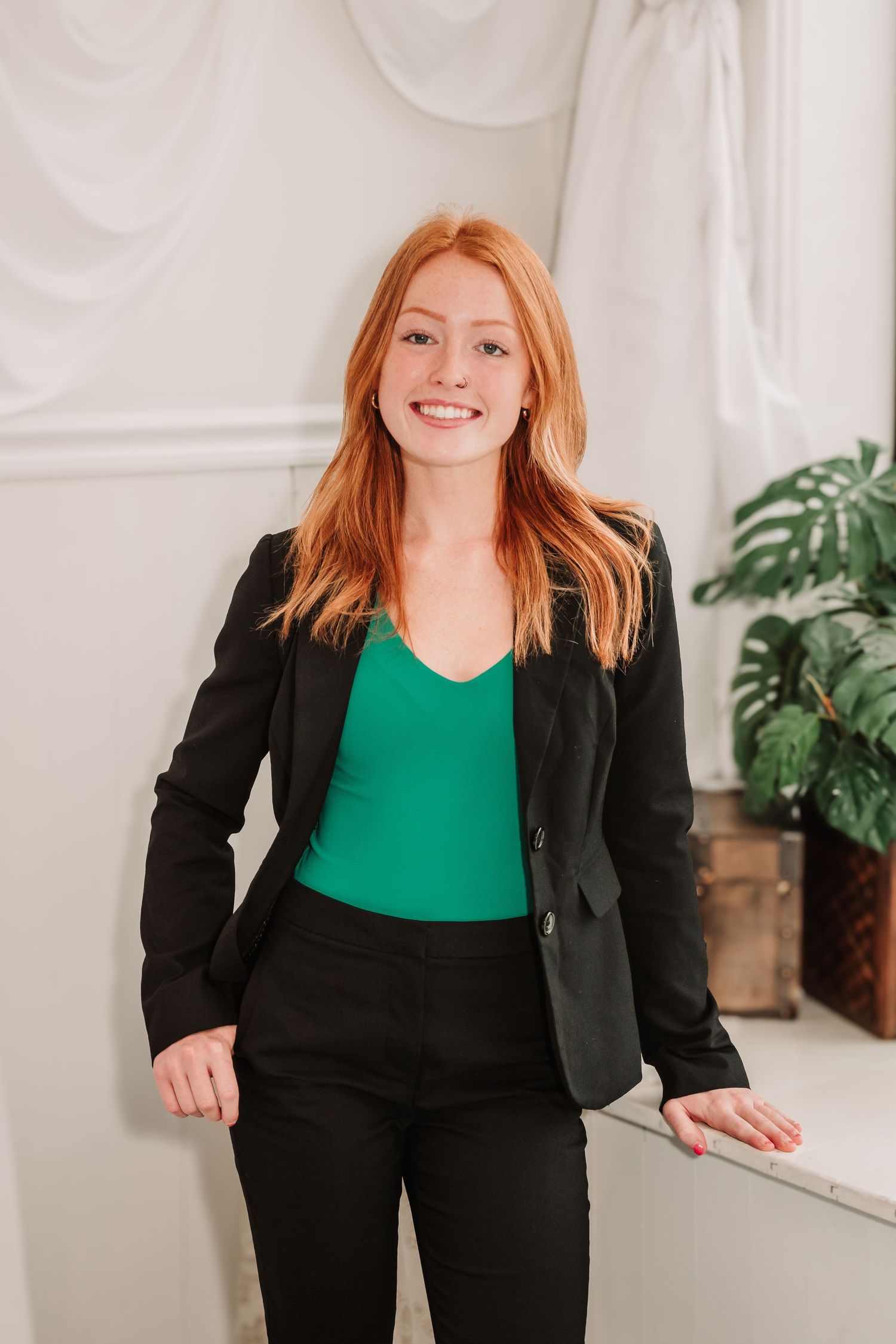 Woman in black blazer and green top poses in professional business attire against white backdrop with plant.