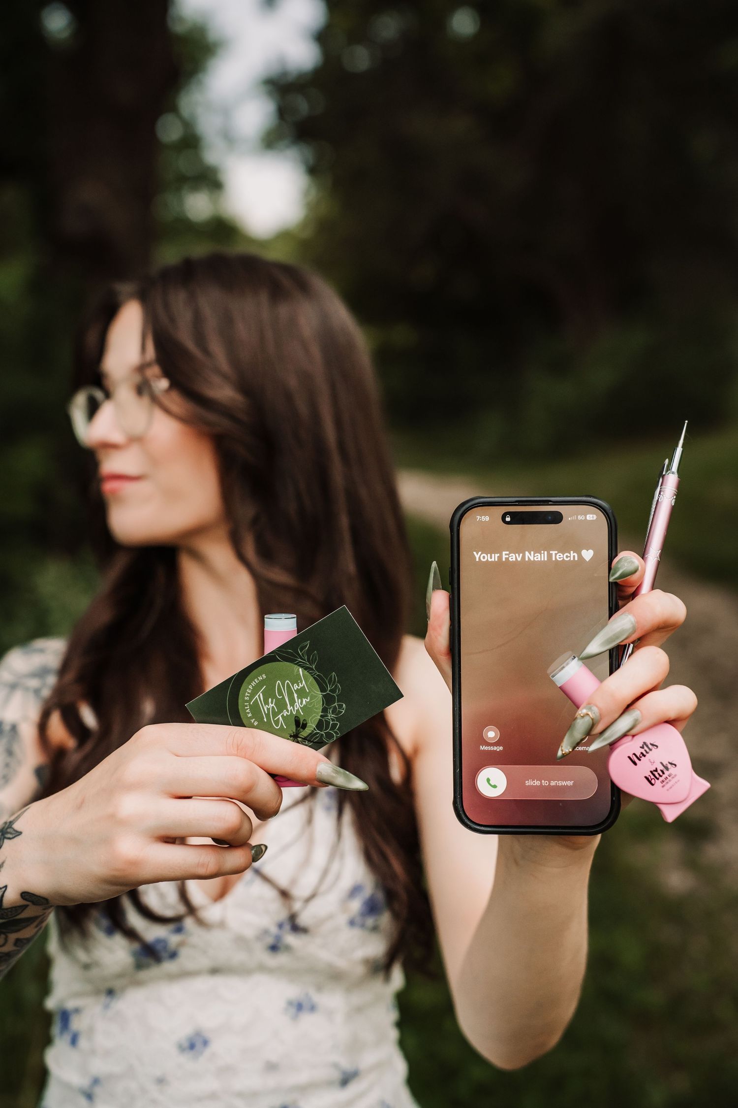Woman holding phone showing nail tech contact and business card outdoors, displaying long decorated nails.