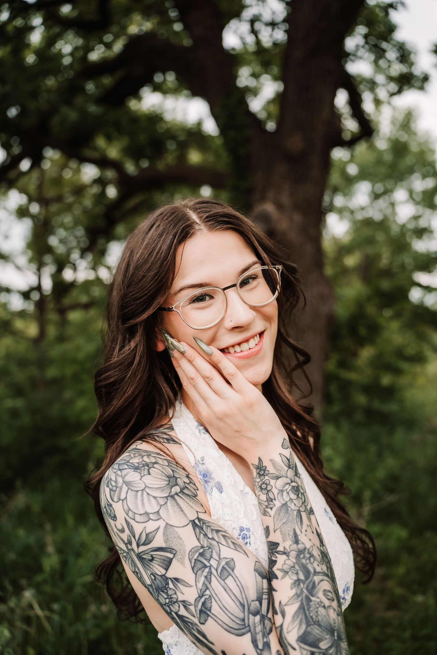 Smiling young woman with glasses and floral arm tattoos posing outdoors near a large tree.