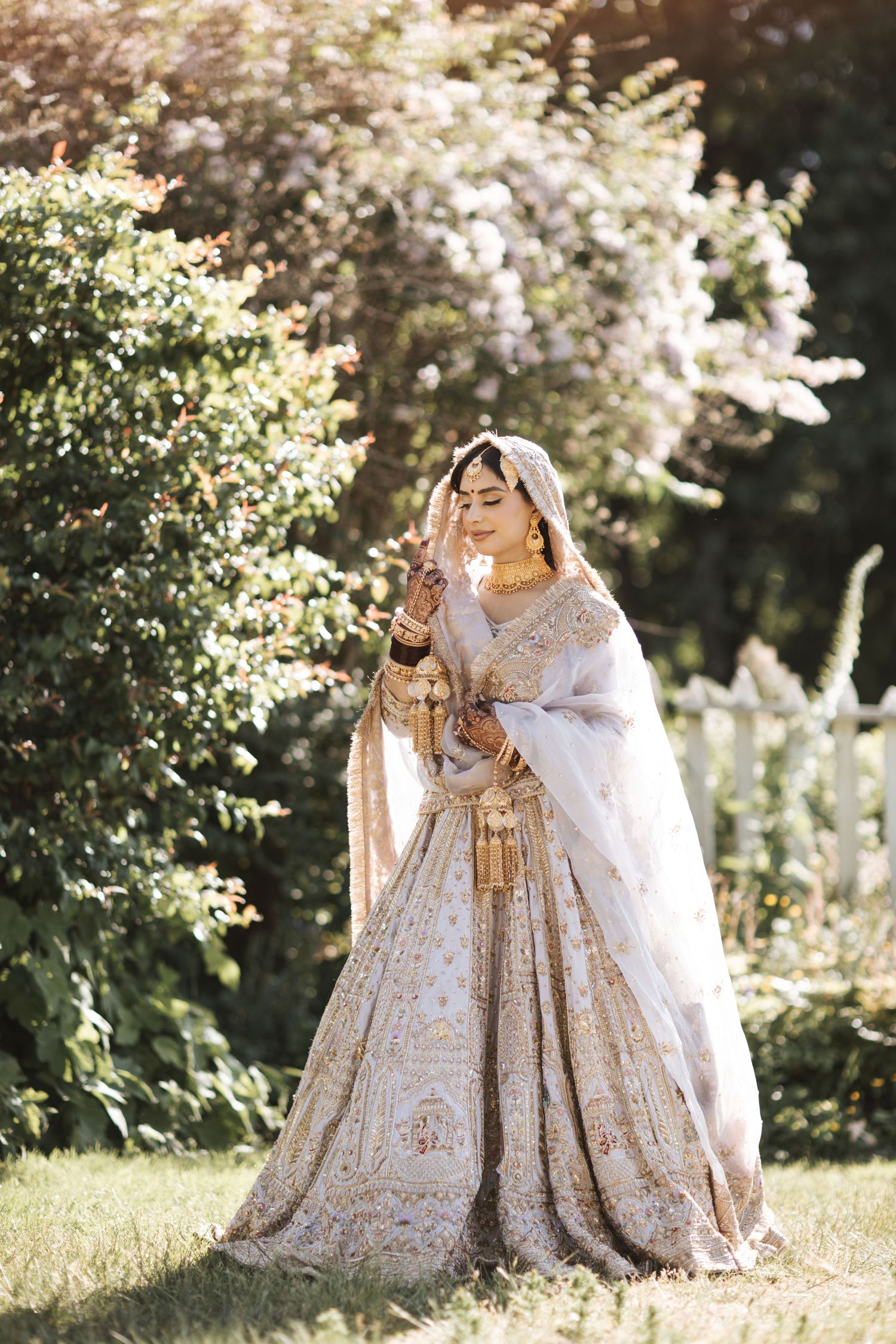 A bride in a white and gold embroidered lehenga with dupatta poses gracefully outdoors among lush green trees.