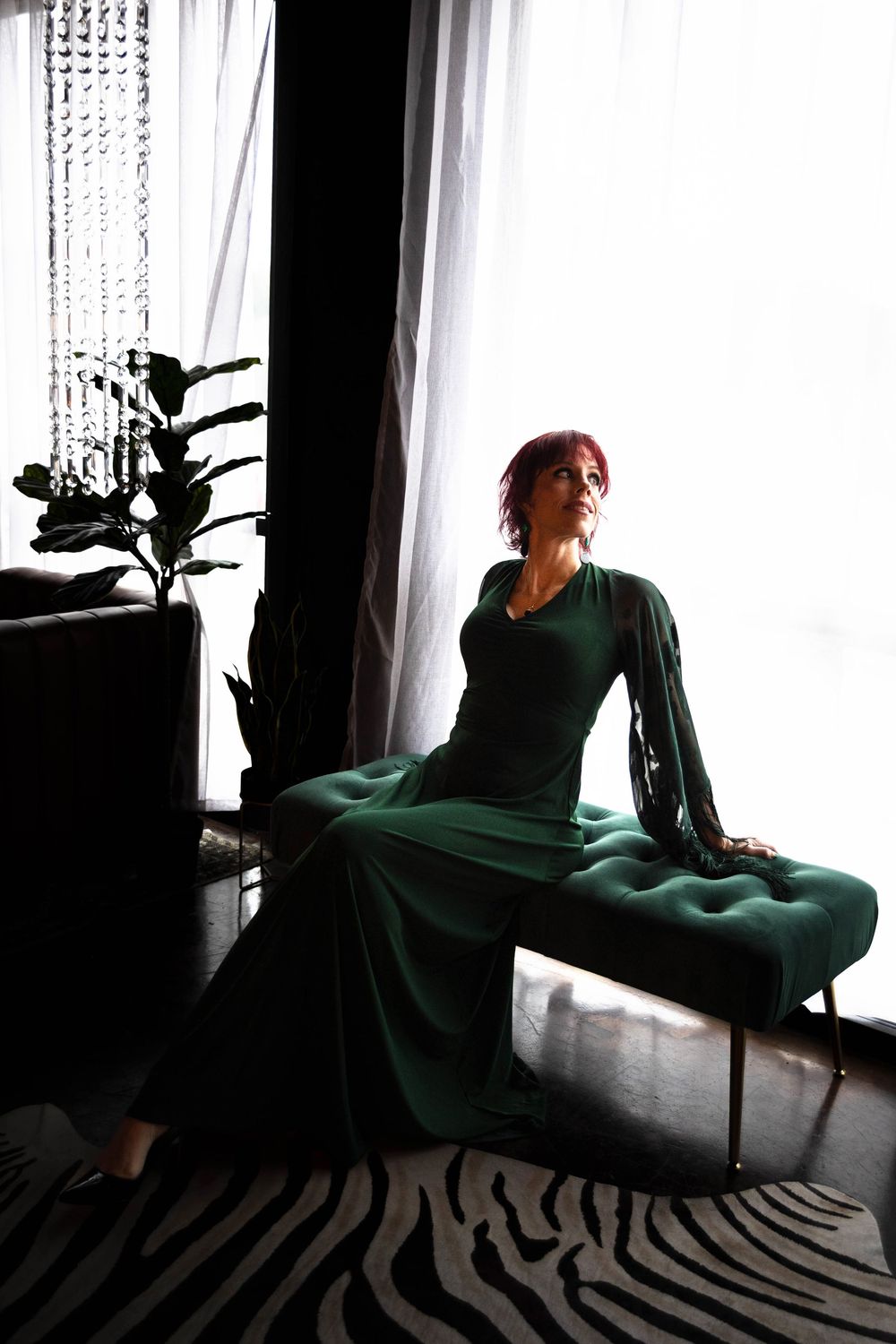 A woman in a green dress poses on a green bench in front of a large window.