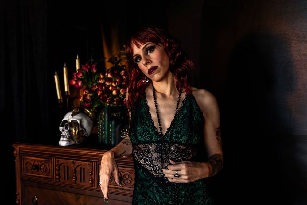 A woman with red hair poses next to a dresser with spooky decor. She looks mysterious.