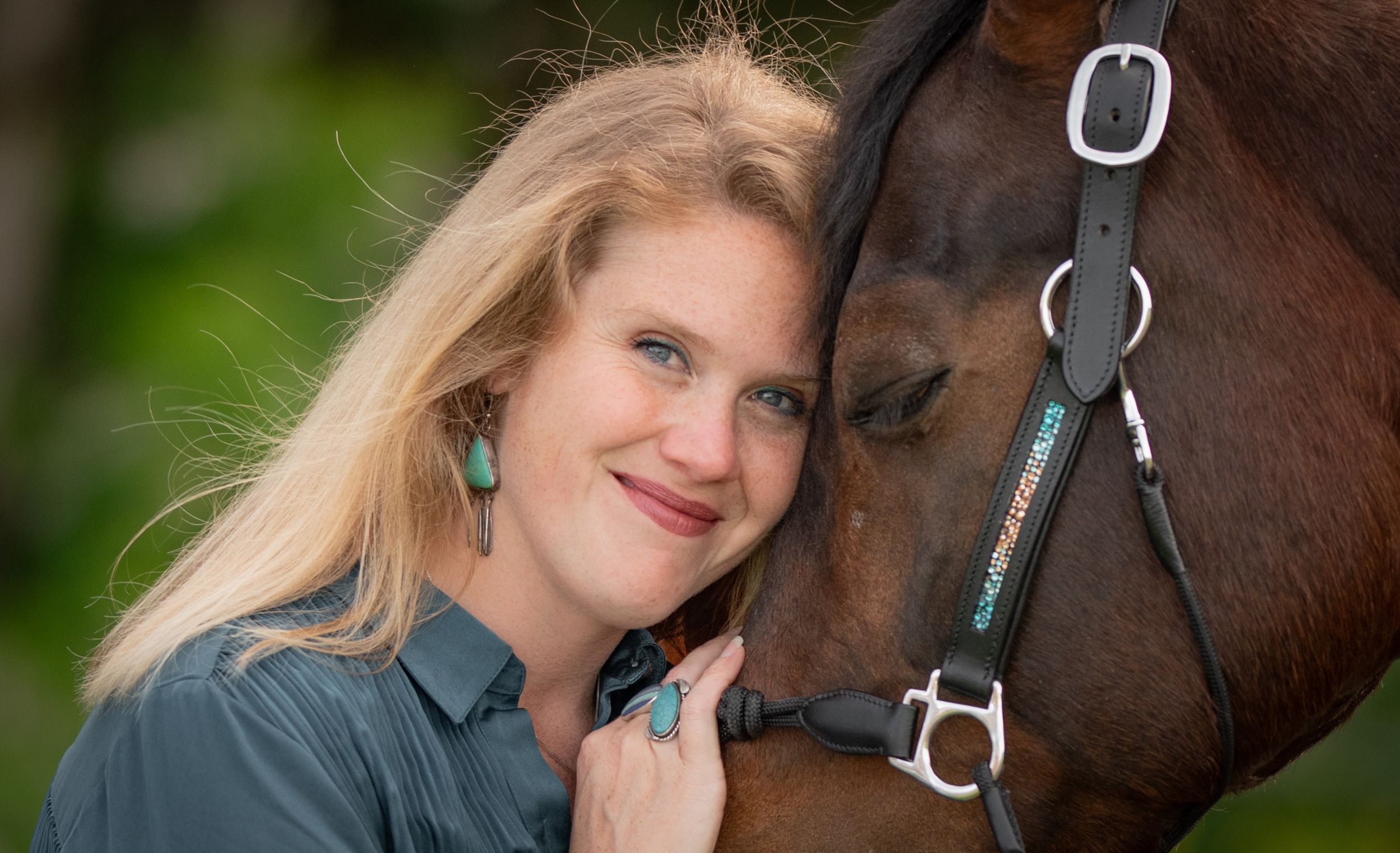 Smiling blonde woman with turquoise earrings poses cheek-to-cheek with a brown horse outdoors.