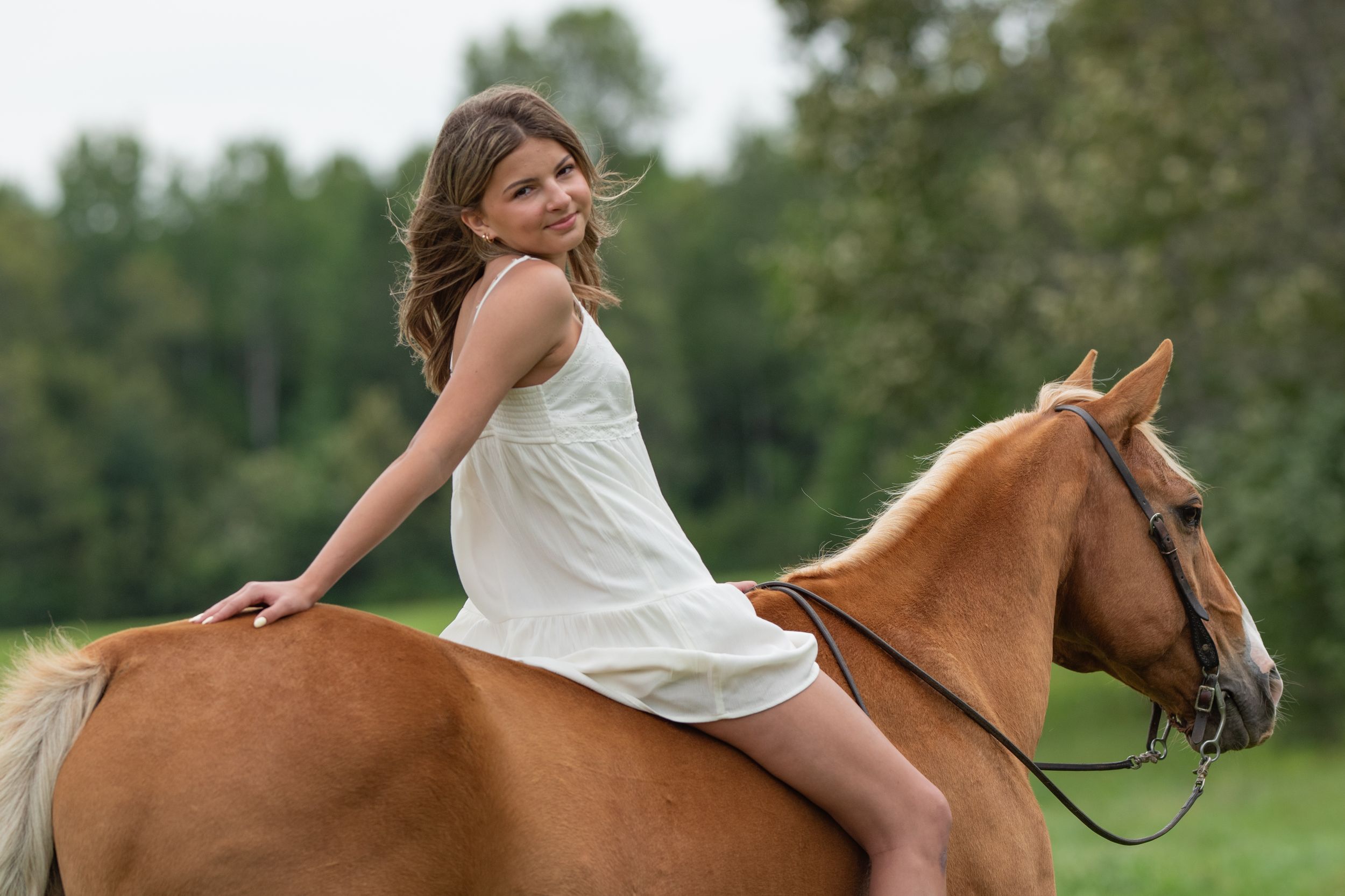 Young girl in white dress rides chestnut horse bareback in lush green field, smiling over shoulder.