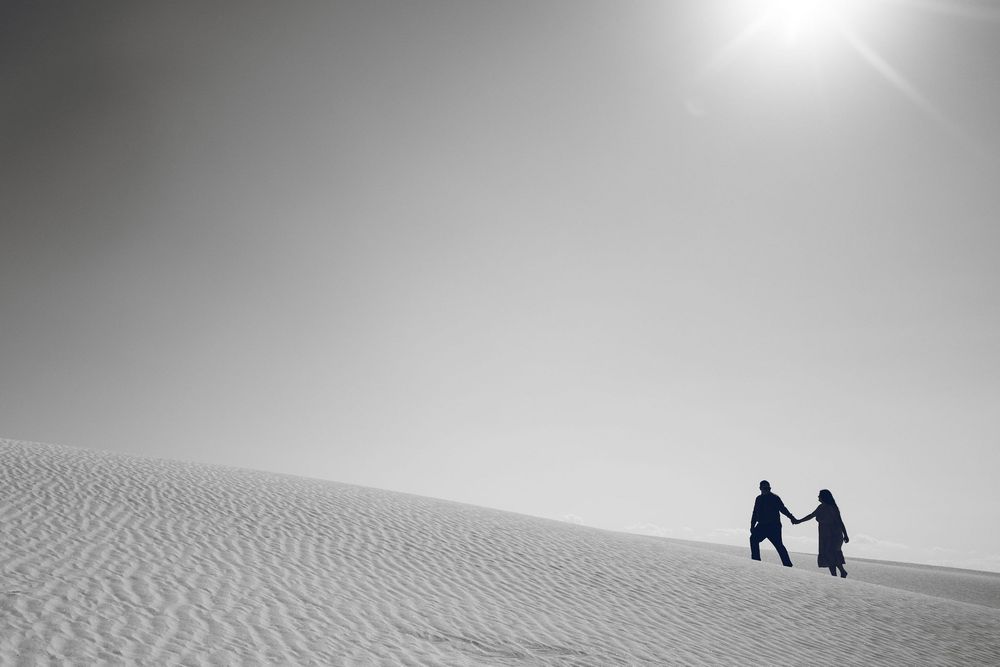 Two people hold hands while walking across white sand dunes under a bright sun in a black and white desert landscape.