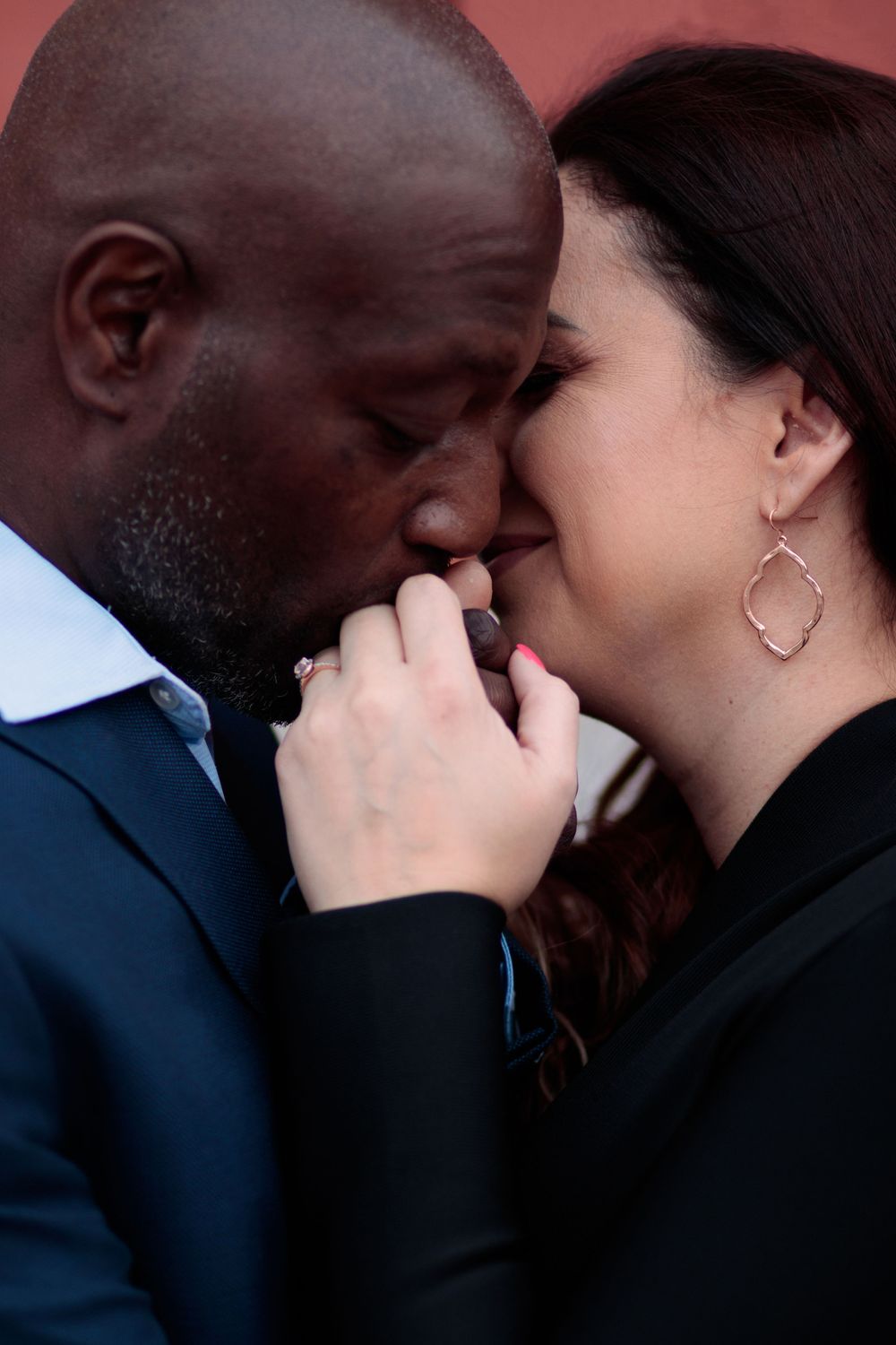 Romantic interracial couple sharing an intimate moment, man kissing woman's hand tenderly against a red wall.