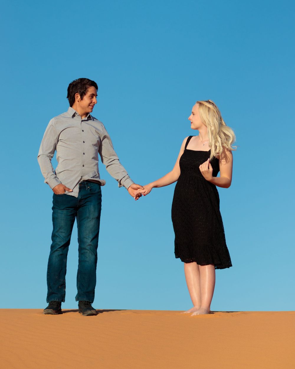 Couple holding hands on sandy desert dunes under clear blue sky, man in jeans and woman in black dress.