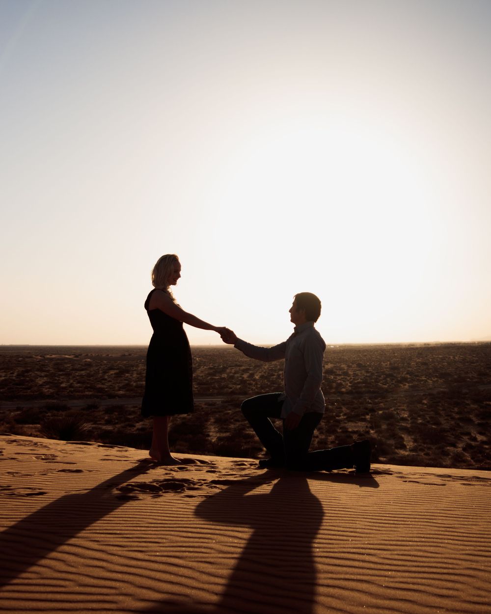 Silhouette of a man proposing on one knee to a woman on a sand dune at sunset in the desert.