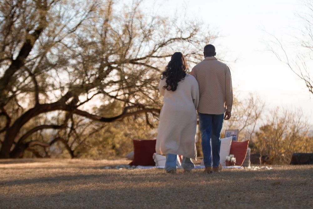 Couple standing together outdoors near bare trees at sunset, viewed from behind in warm winter coats.