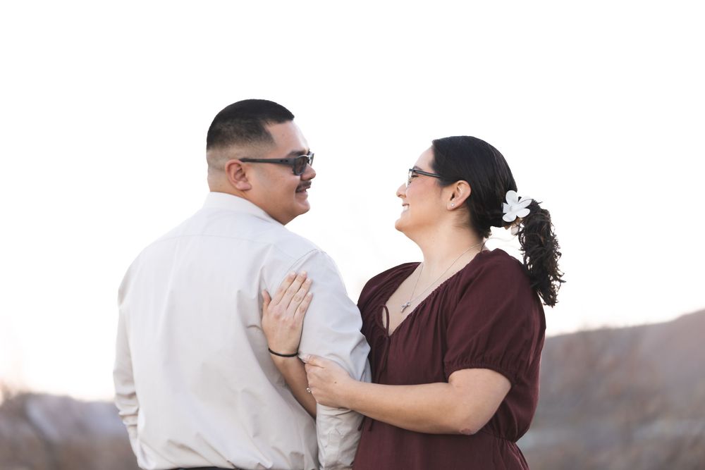 Couple smiling at each other outdoors, woman with flower in hair wearing burgundy dress, man in white shirt with sunglasses.