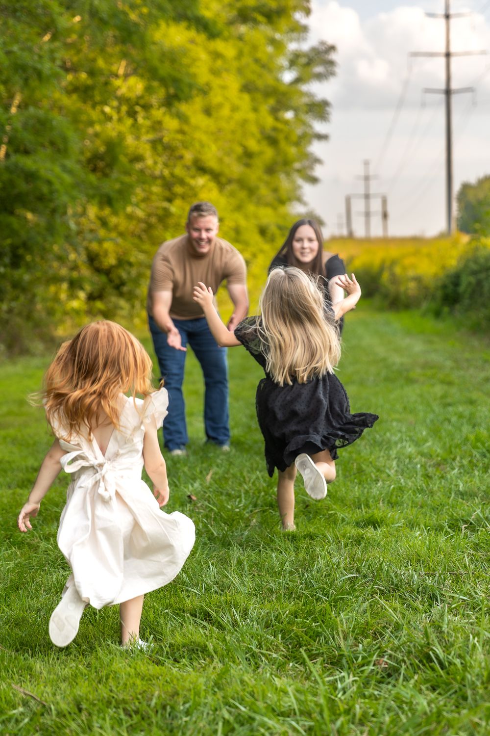 Children in dresses run playfully through green grass path towards adults.