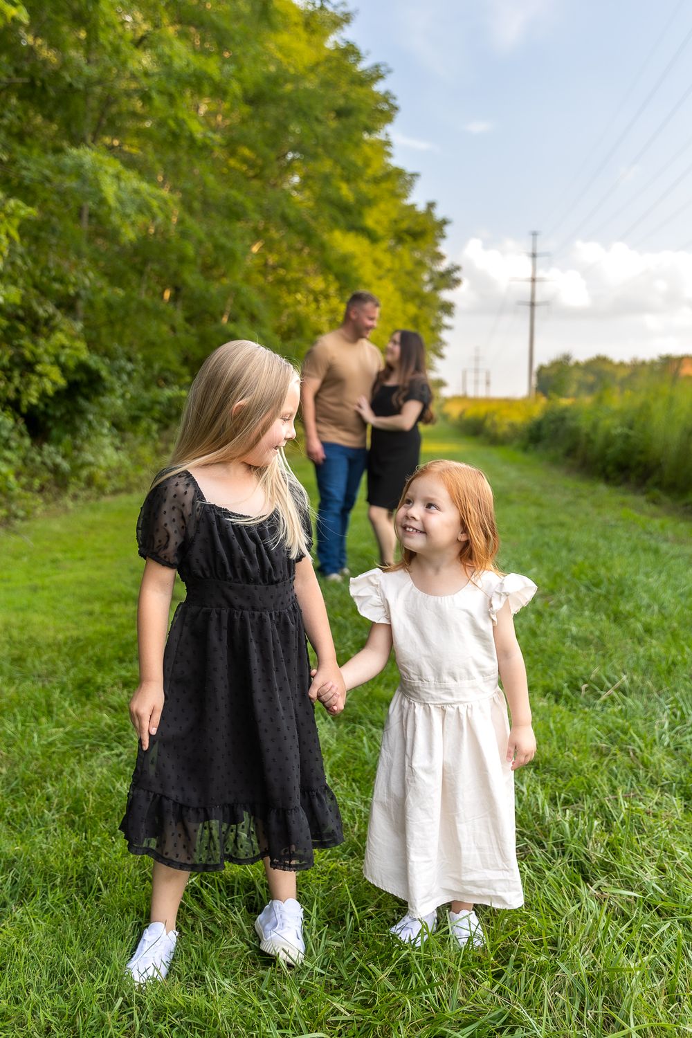 Two young children in dresses share sweet moment on grassy path with trees.