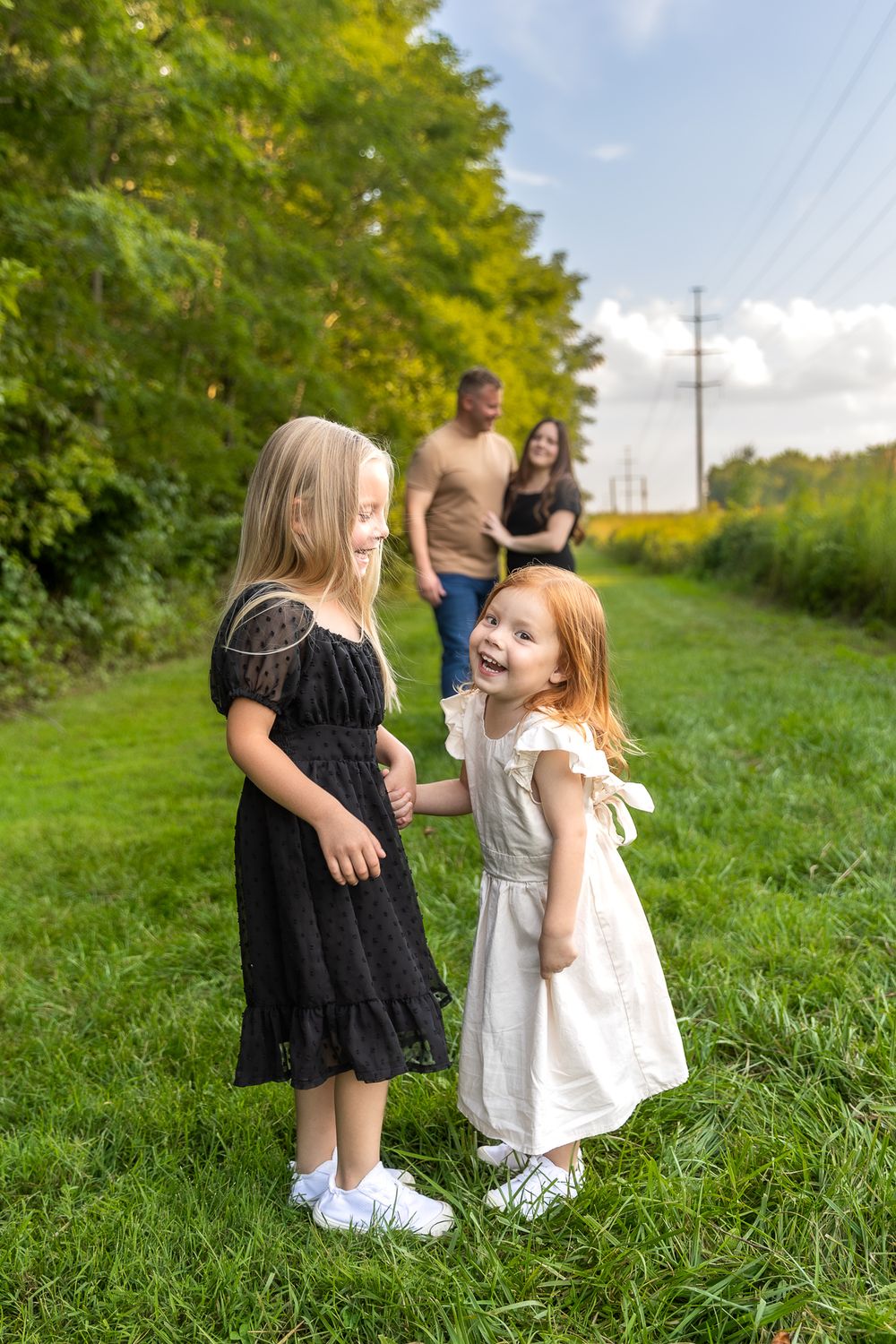 Two children in contrasting dresses share laughs on grassy path with adults in background.
