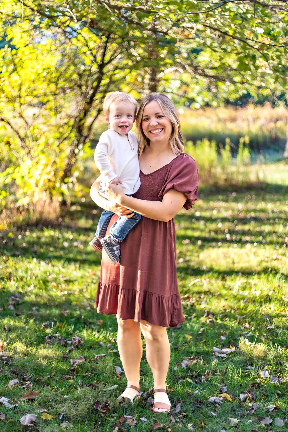 Woman in burgundy dress holding child while posing under trees in golden autumn sunlight.