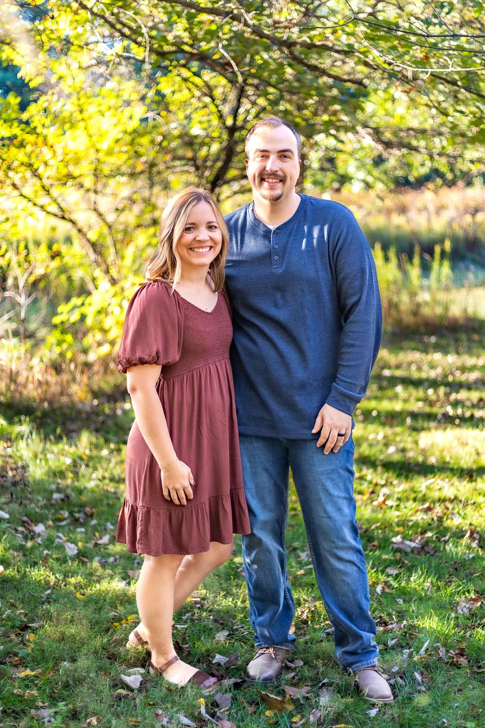 Couple standing together outdoors in casual attire against autumn trees and natural lighting.