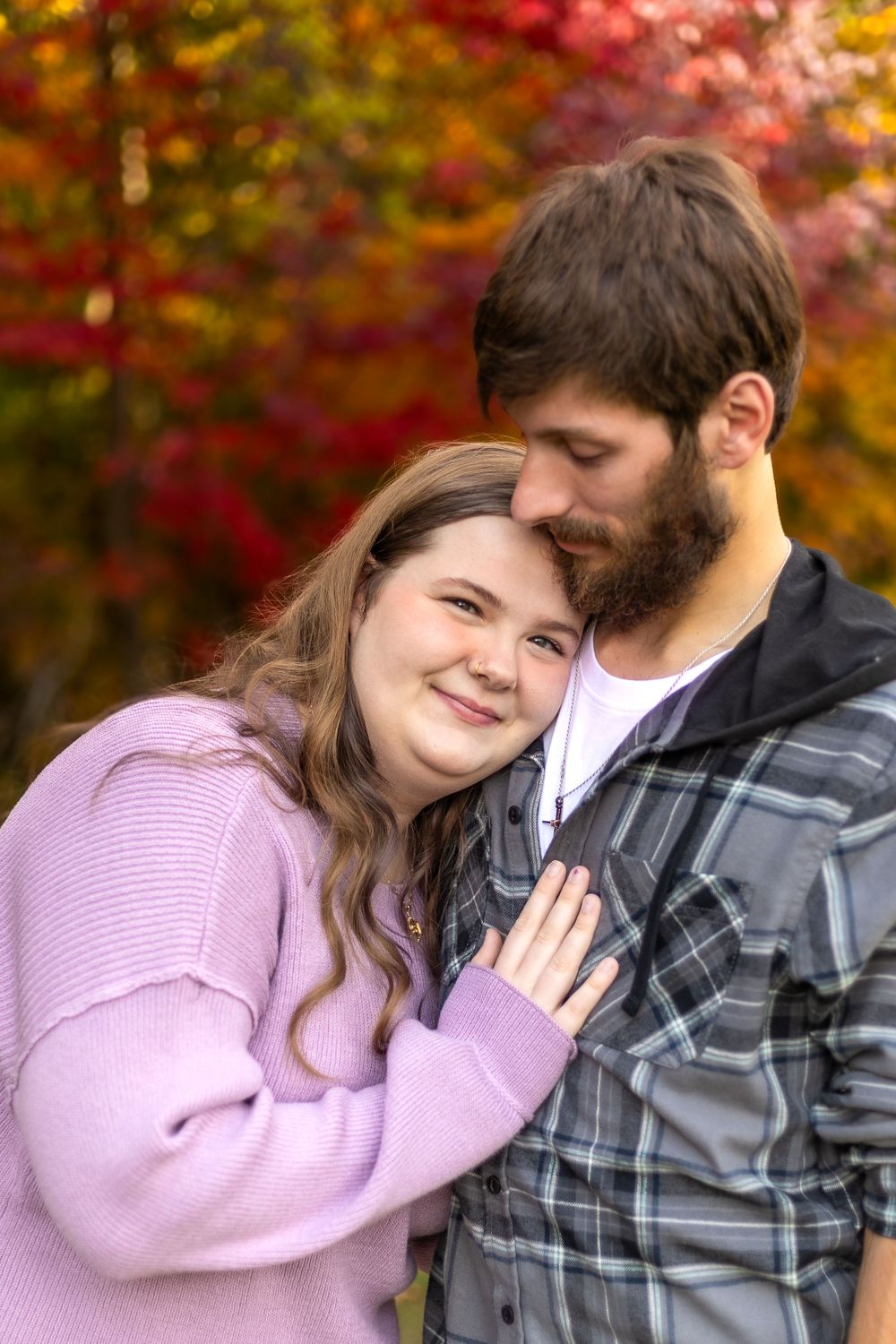 A couple shares an intimate moment against vibrant fall foliage in casual outdoor wear.