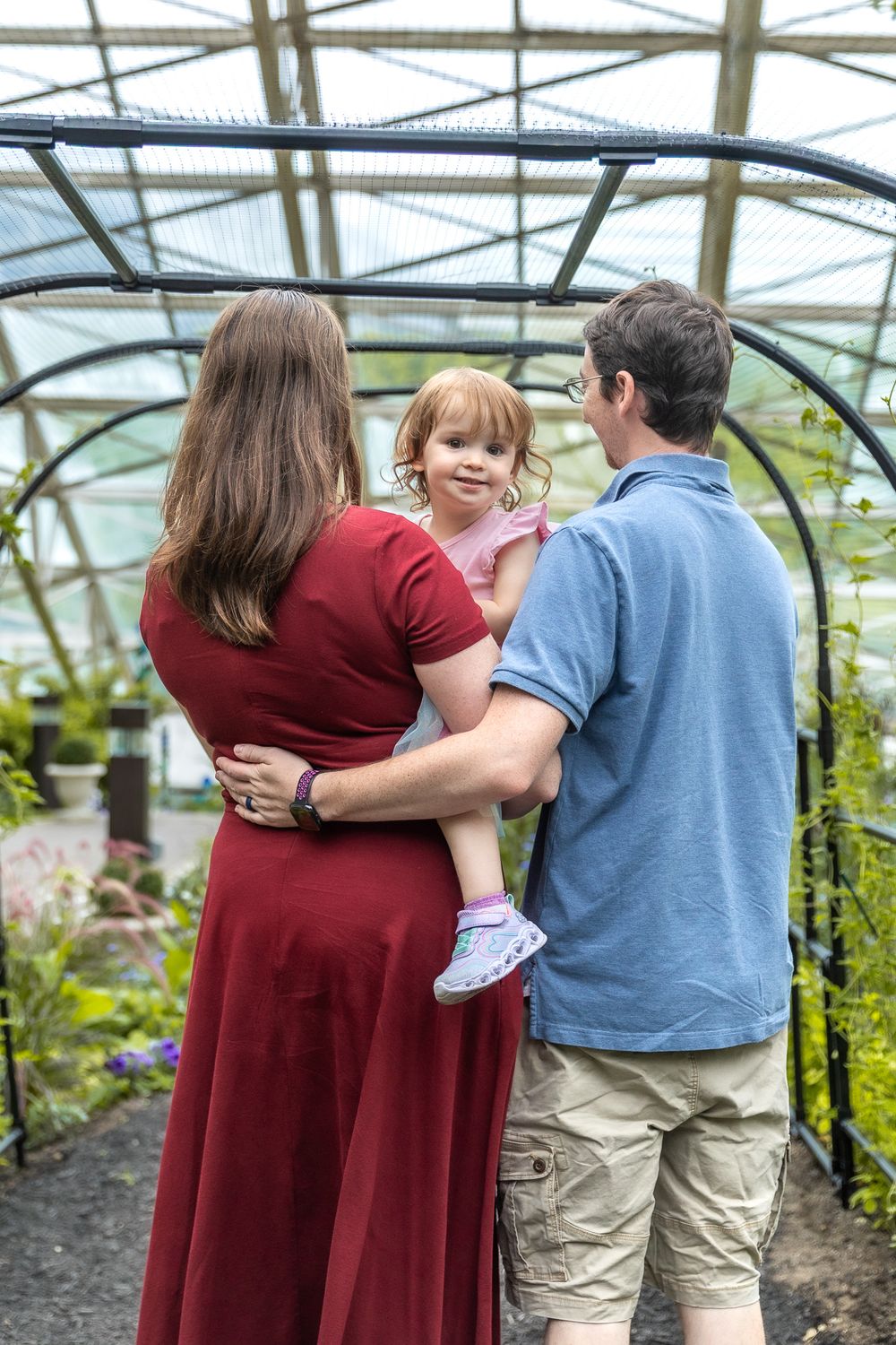 Couple embraces and laughs together in a greenhouse setting with glass roof and plants visible in background.