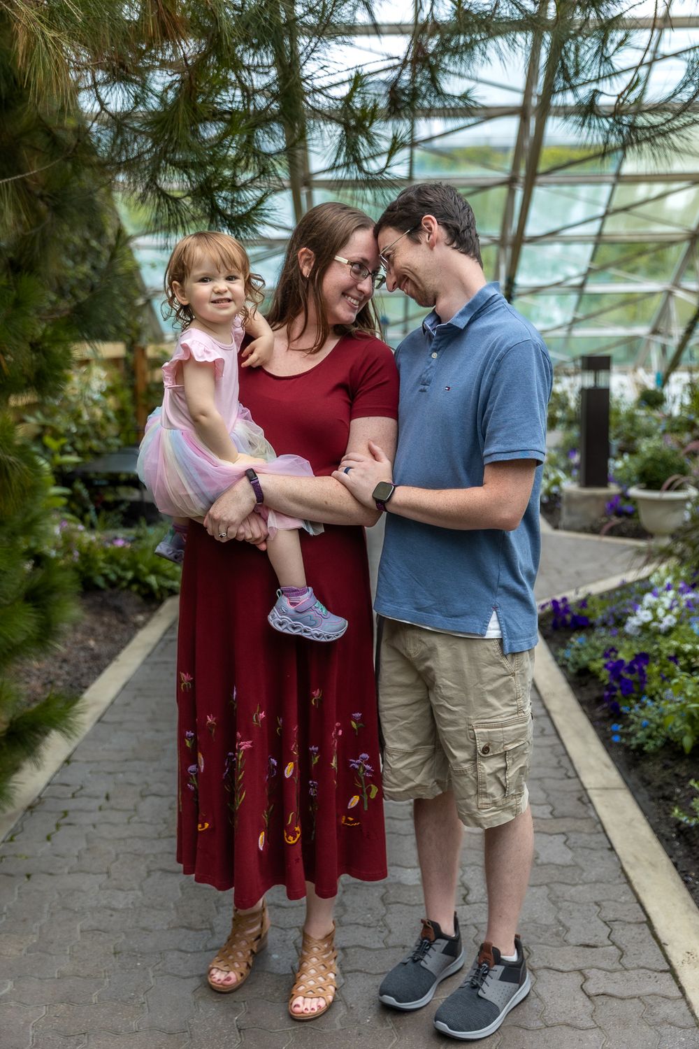 Family wearing burgundy and casual clothing poses together in a greenhouse garden setting with flowering plants.