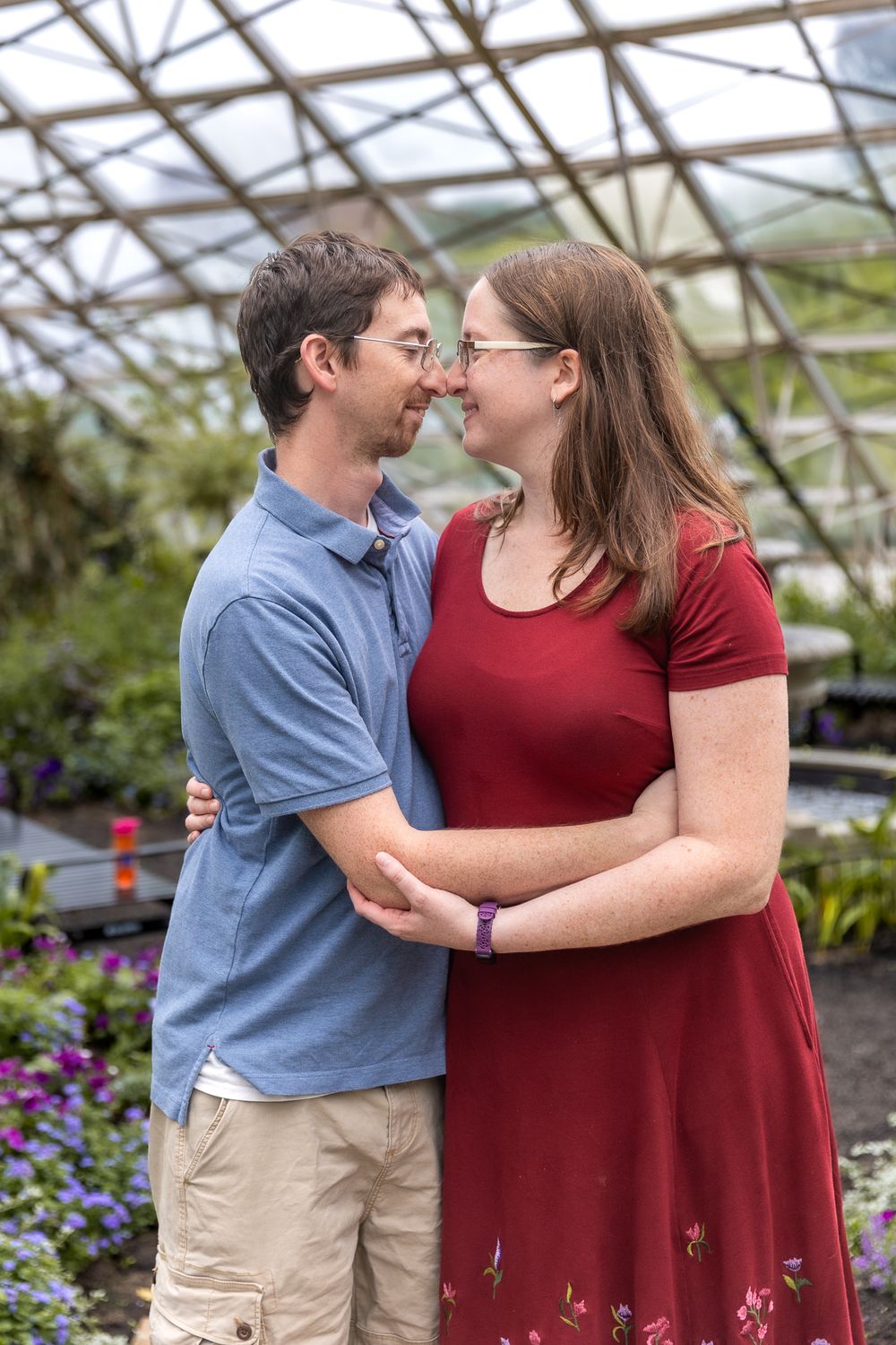 Couple shares romantic moment together while standing in a greenhouse surrounded by colorful flowers and plants.