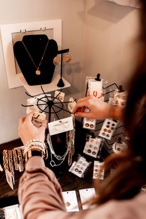 Hands arranging jewelry on a display stand with necklaces, bracelets and earrings for sale on a white surface.