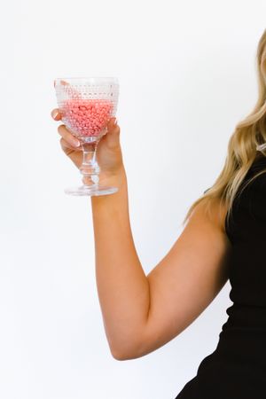 Woman in black dress holding a crystal goblet filled with pink candy beads against white background.