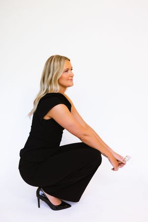 Blonde woman in black dress and heels crouching gracefully against a white background.