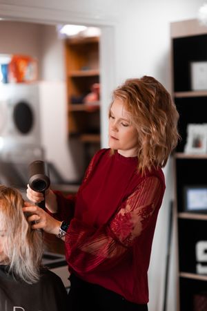 Professional hairstylist wearing a red lace-sleeve sweater uses styling tools in a salon setting.