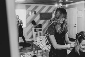 A stylist works on client's hair in a modern salon with striped wallpaper while another person stands in background.