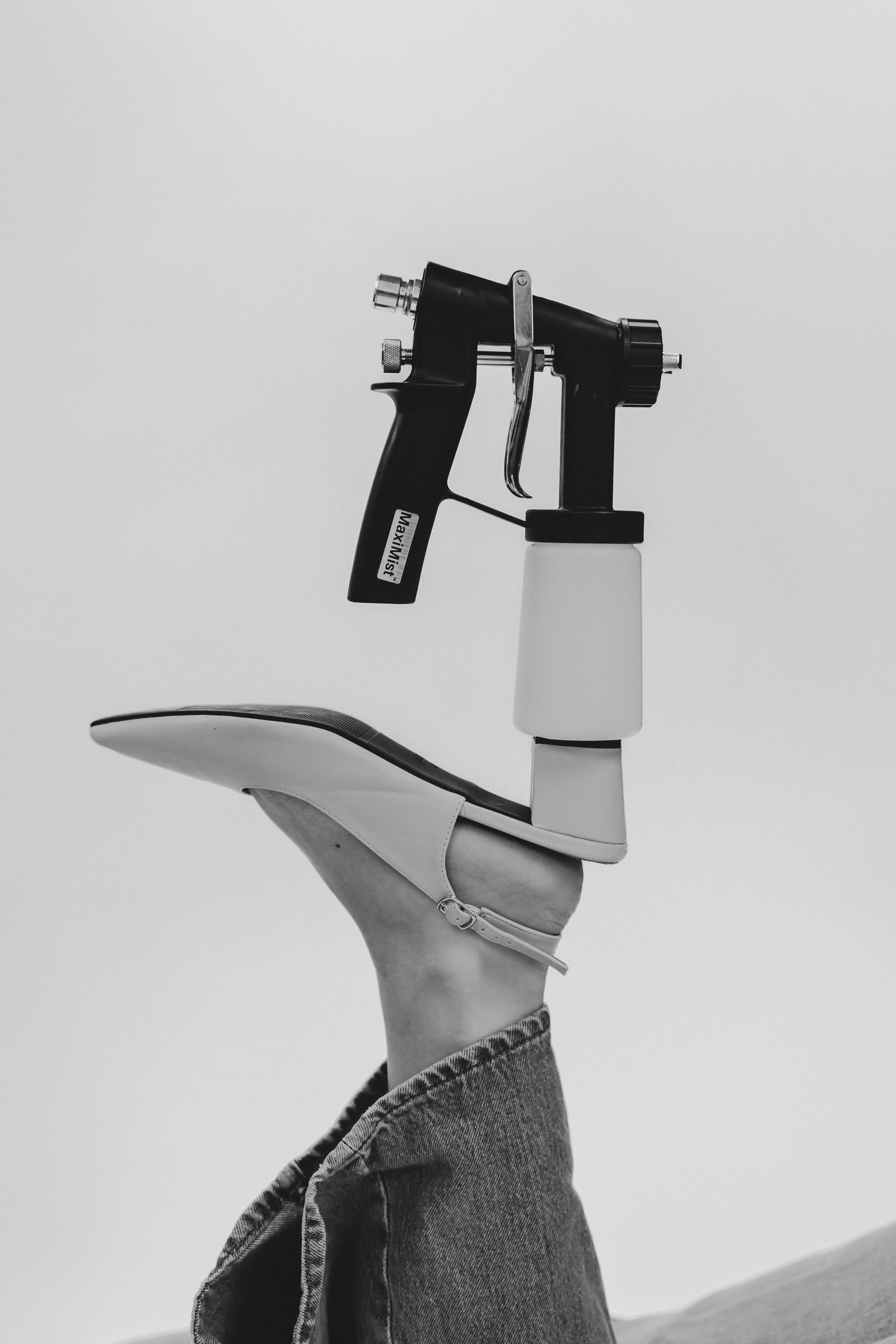 Black and white photo of a leg in jeans and heels balancing a spray gun on the foot against a light background.