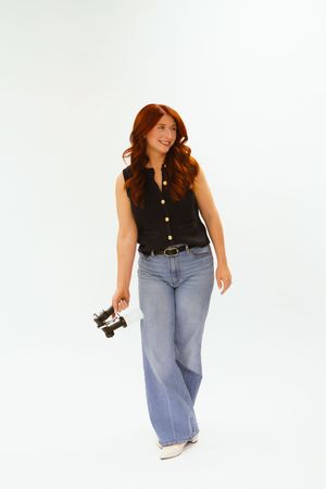 A woman with red hair in a black sleeveless top and wide-leg jeans holds a vintage camera while walking against a white background.