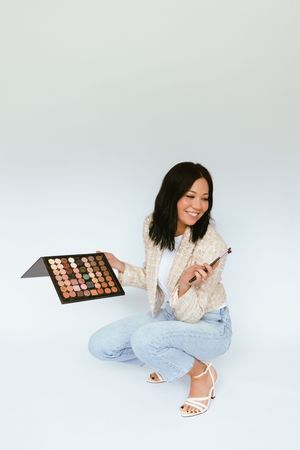 A person in casual attire displays a makeup palette while sitting against a white backdrop.