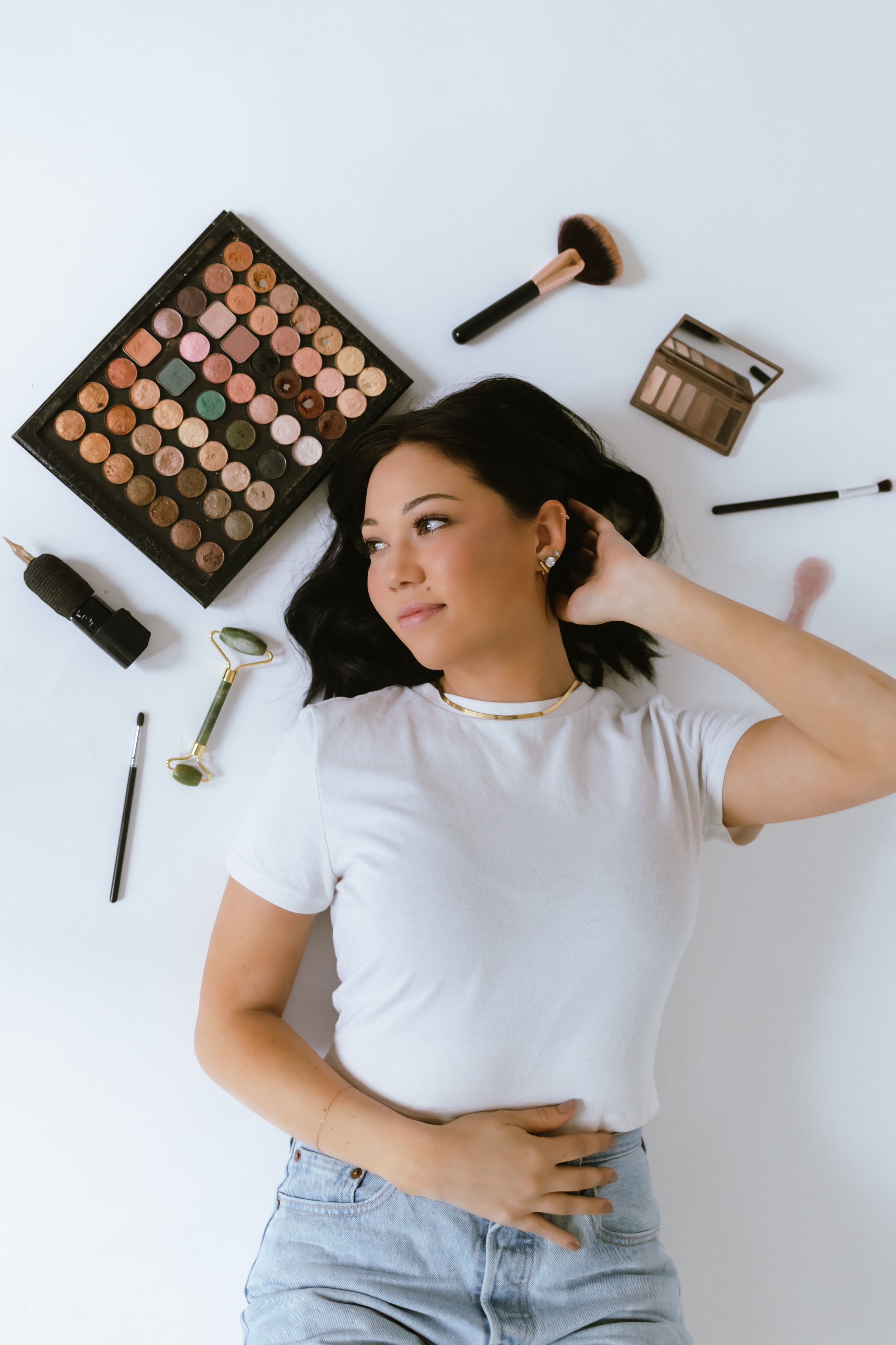 Makeup products and brushes arranged around a person lying on a white background.