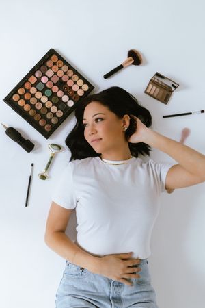 Makeup products and brushes arranged around a person lying on a white background.