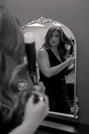 A black and white artistic photograph shows someone's reflection in an ornate mirror posing in dark attire.