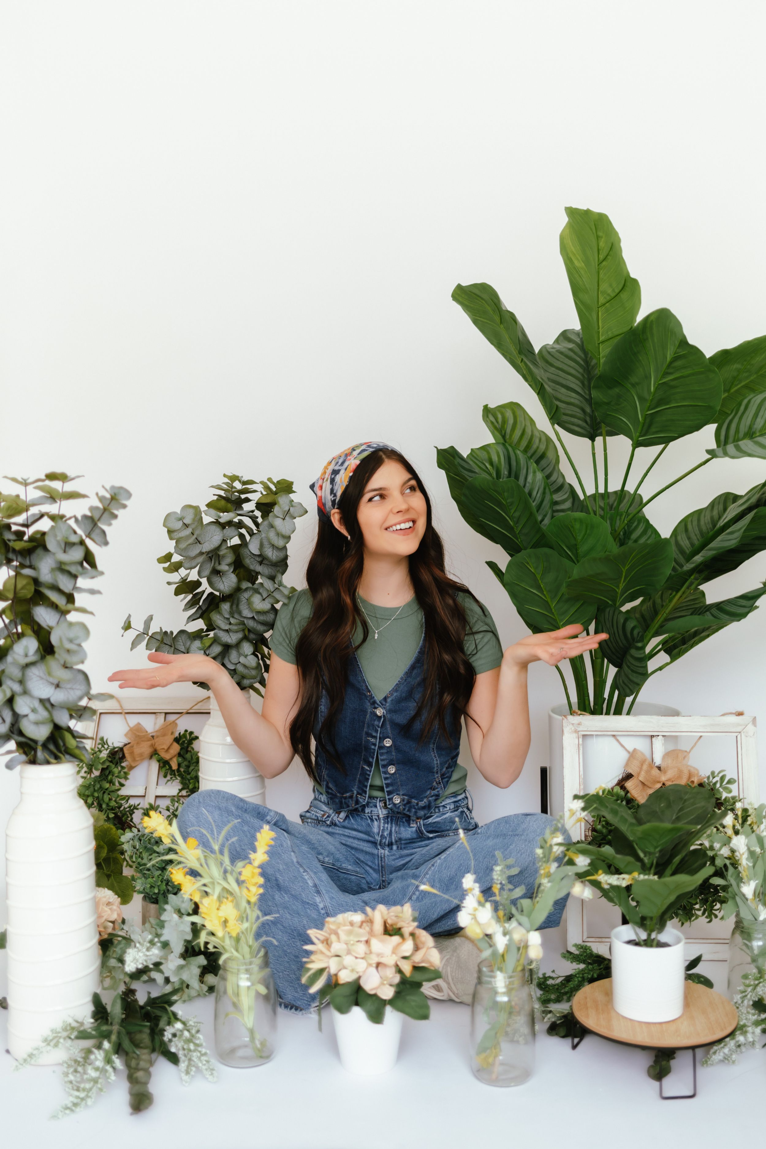 Person in denim sits on white floor surrounded by houseplants and floral arrangements in decorative vases.