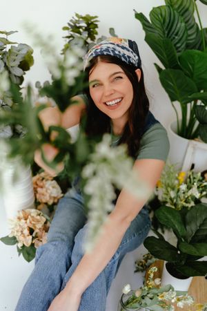 A casual portrait surrounded by lush houseplants and floral arrangements in a bright, airy setting.