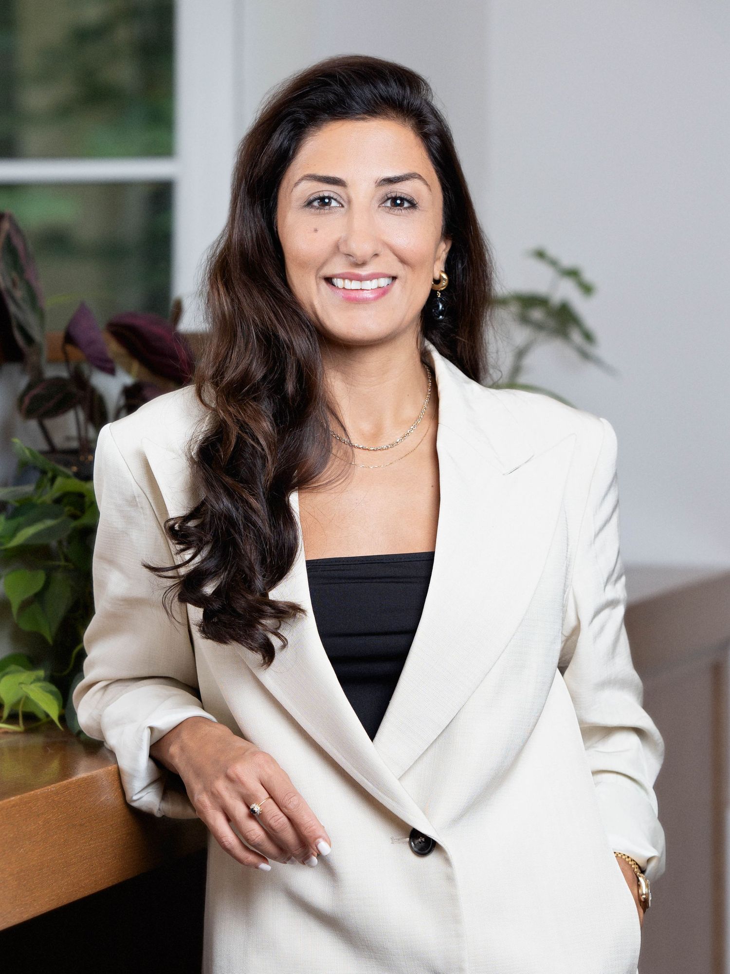 Professional portrait of someone in a crisp white blazer and black top with long dark hair and jewelry against an office setting.