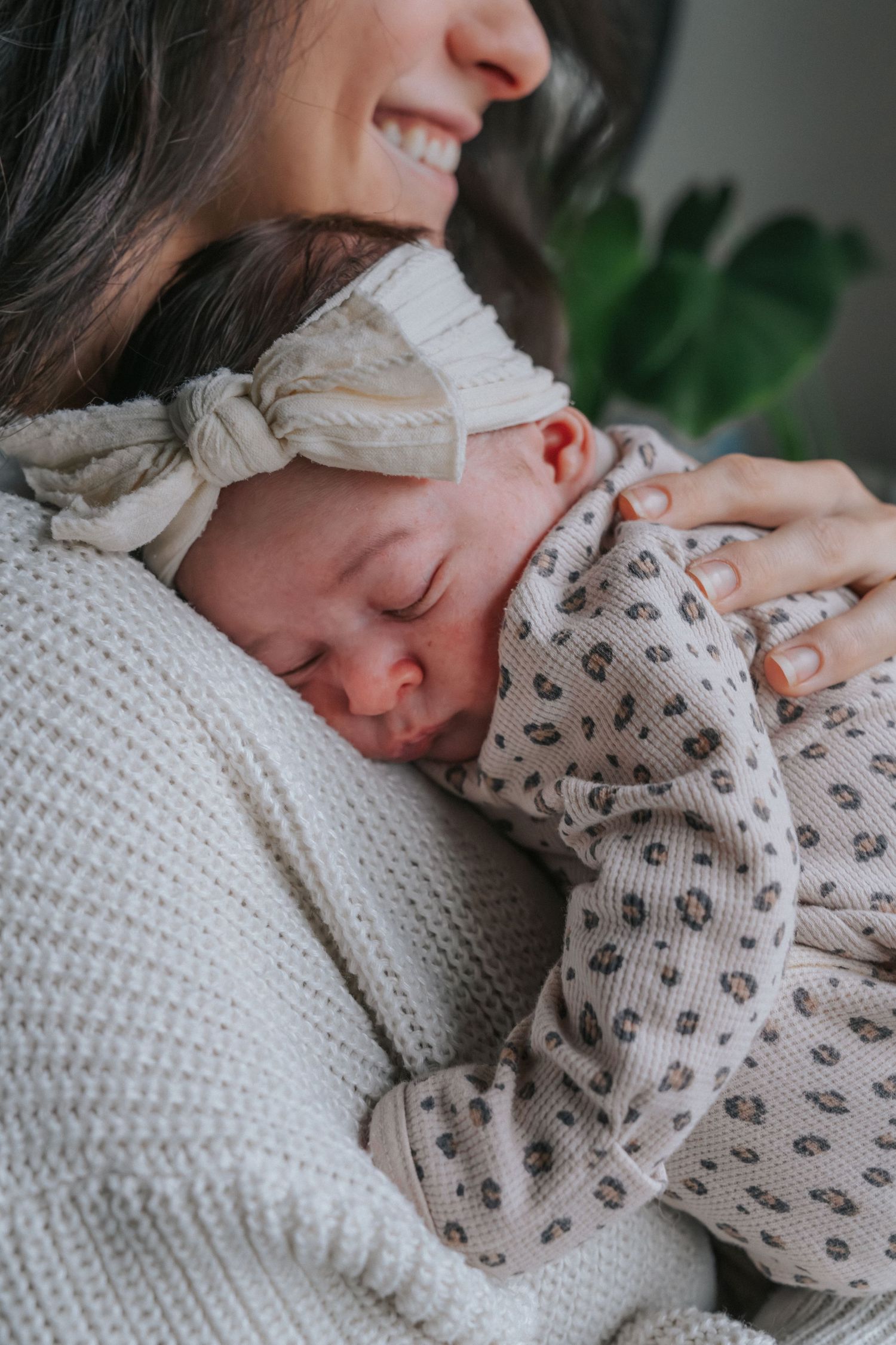 A sleeping newborn baby wearing a patterned onesie and bow headband rests peacefully against her mom on a white knit sweater.