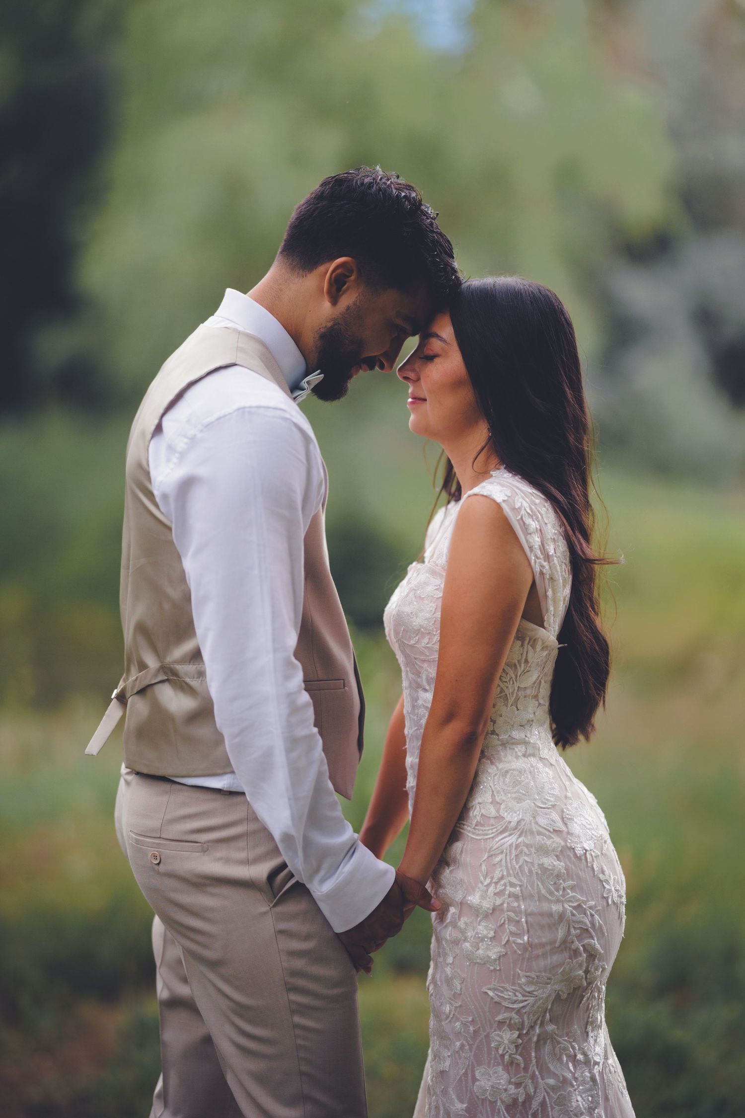 A bride and groom share a quiet, intimate moment, standing close with their foreheads touching outdoors.