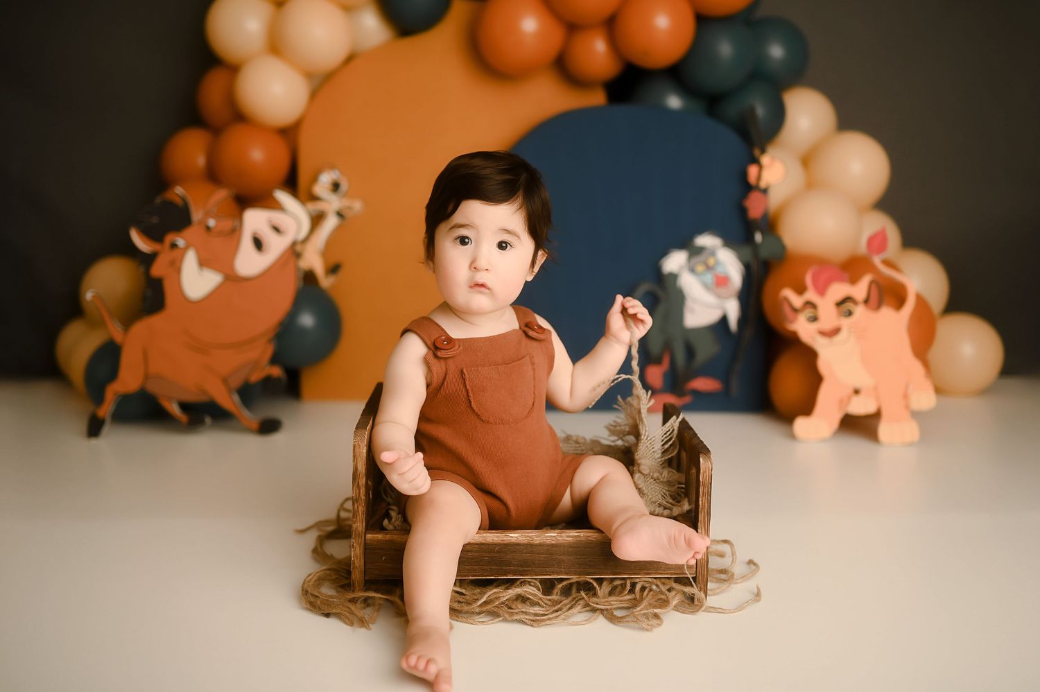 Baby boy in rust romper sits in wooden crate with Lion King themed balloon backdrop for first birthday photo.