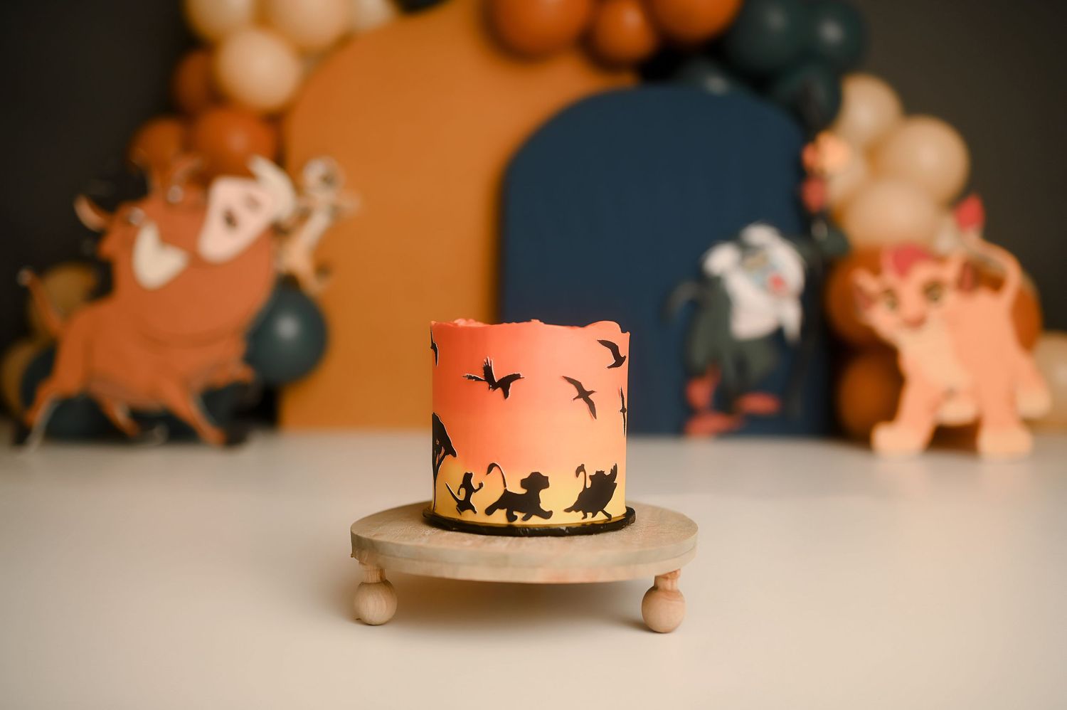 Lion King themed smash cake with silhouette design on orange sunset backdrop with balloon garland.