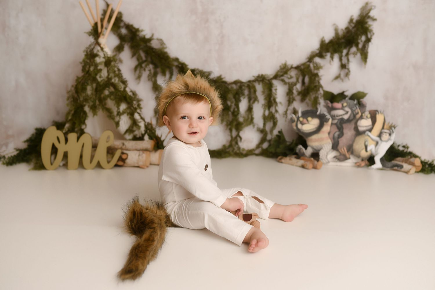Smiling toddler in white onesie with fox tail sits at woodland-themed first birthday session.