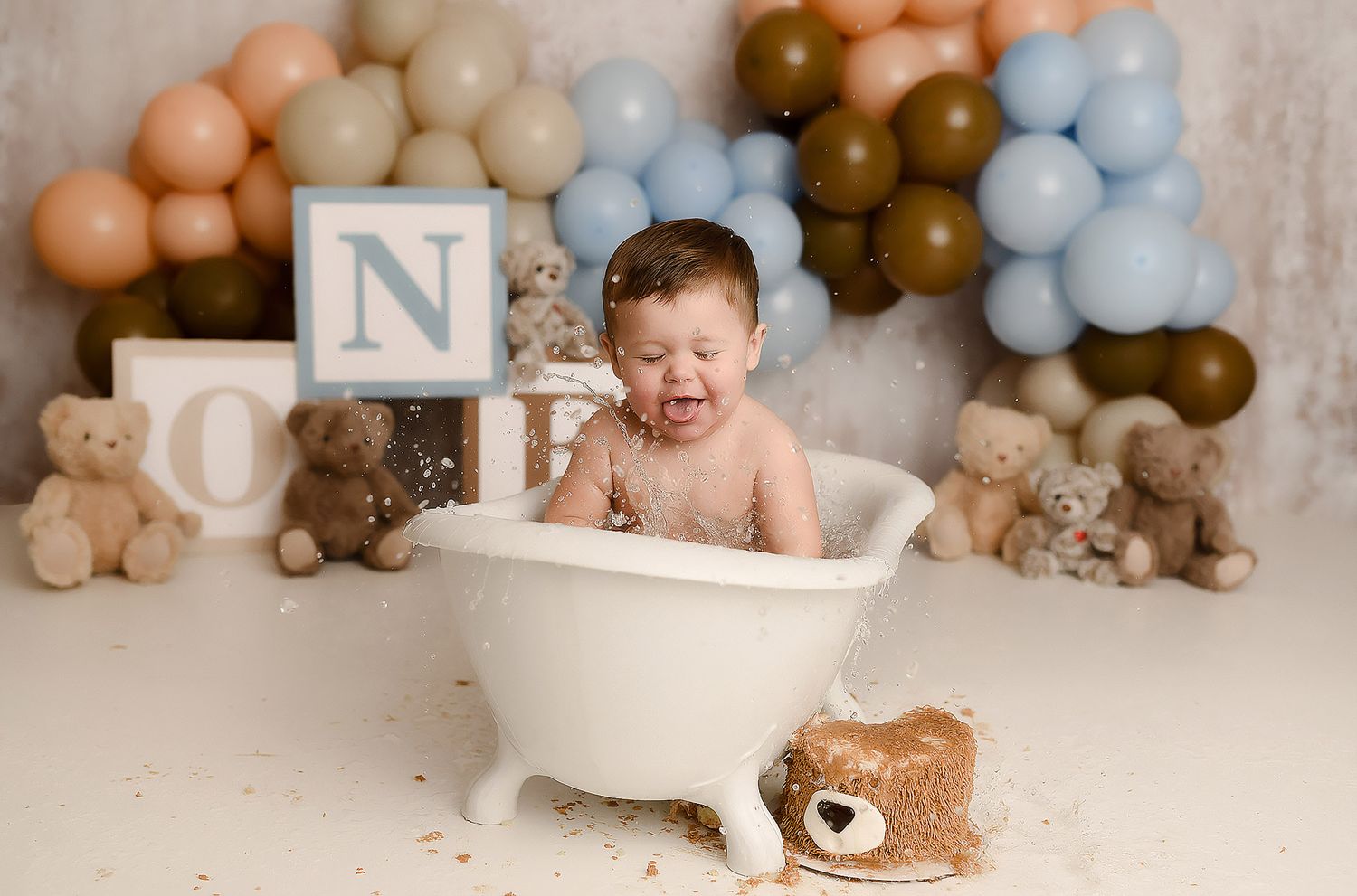 Crying baby sits in white clawfoot tub with teddy bear ONE blocks and blue brown balloon backdrop.