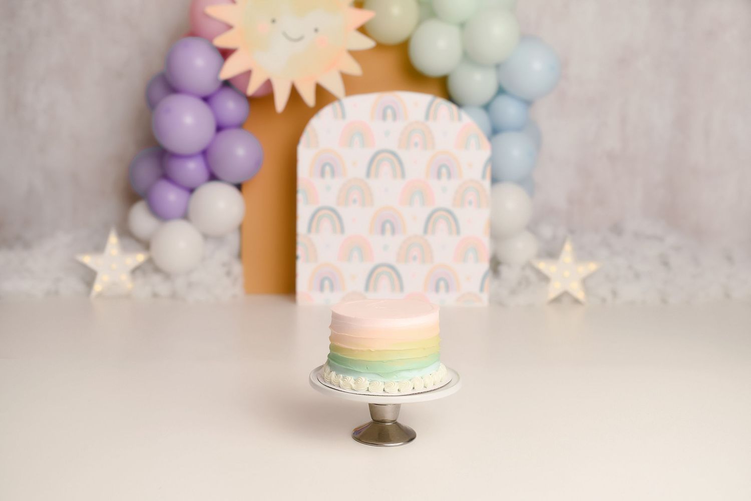 Pastel rainbow-themed first birthday smash cake on stand with colorful balloon arch and sun backdrop.
