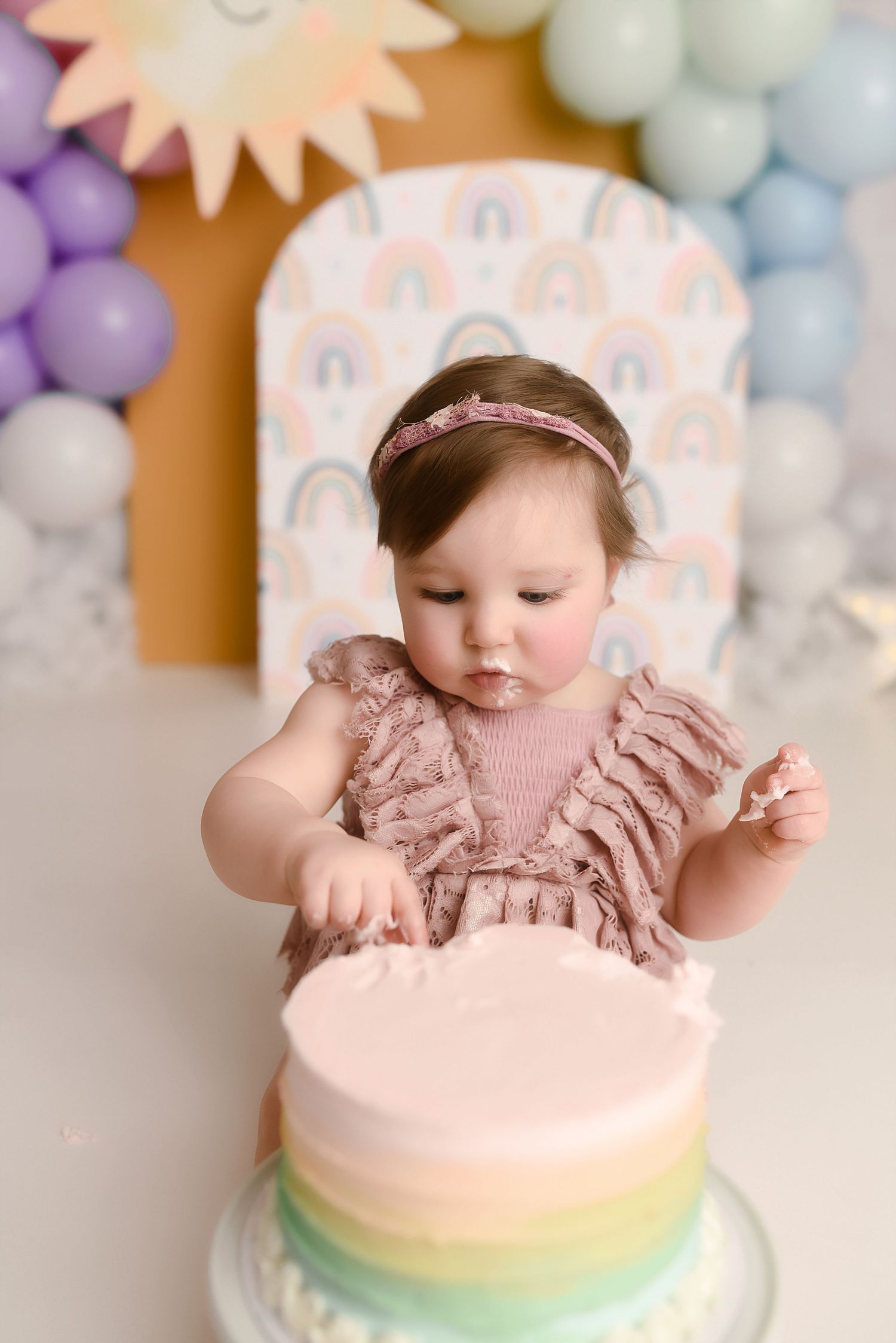 Baby girl in pink dress smashing rainbow birthday cake at colorful balloon backdrop first birthday party.