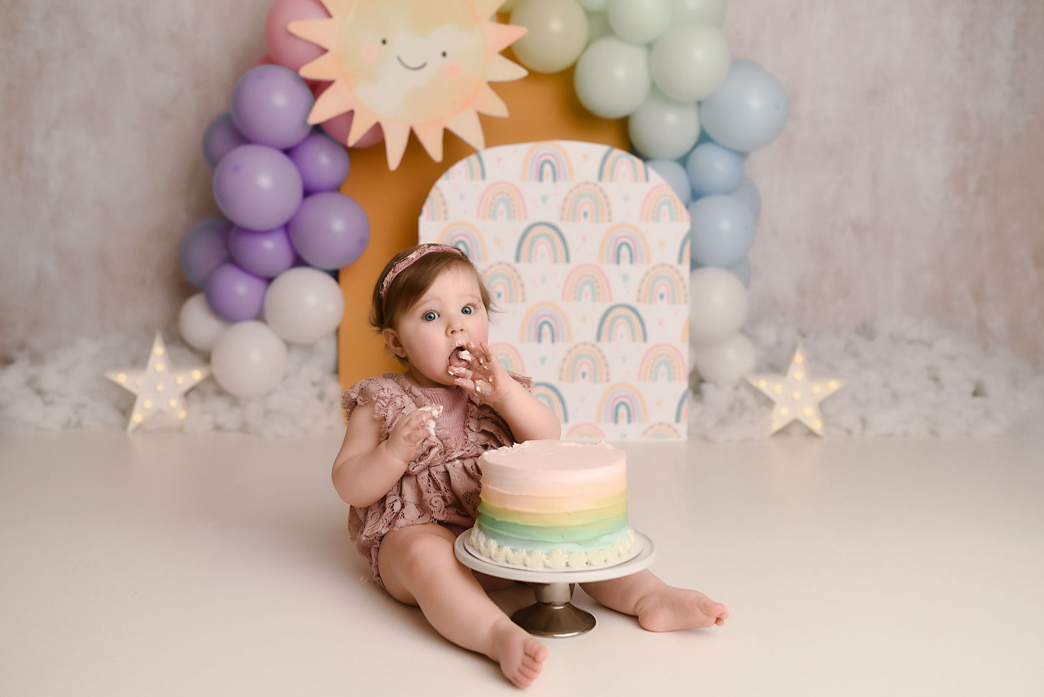 Baby girl eating pastel rainbow first birthday smash cake with colorful balloon arch and sun backdrop.