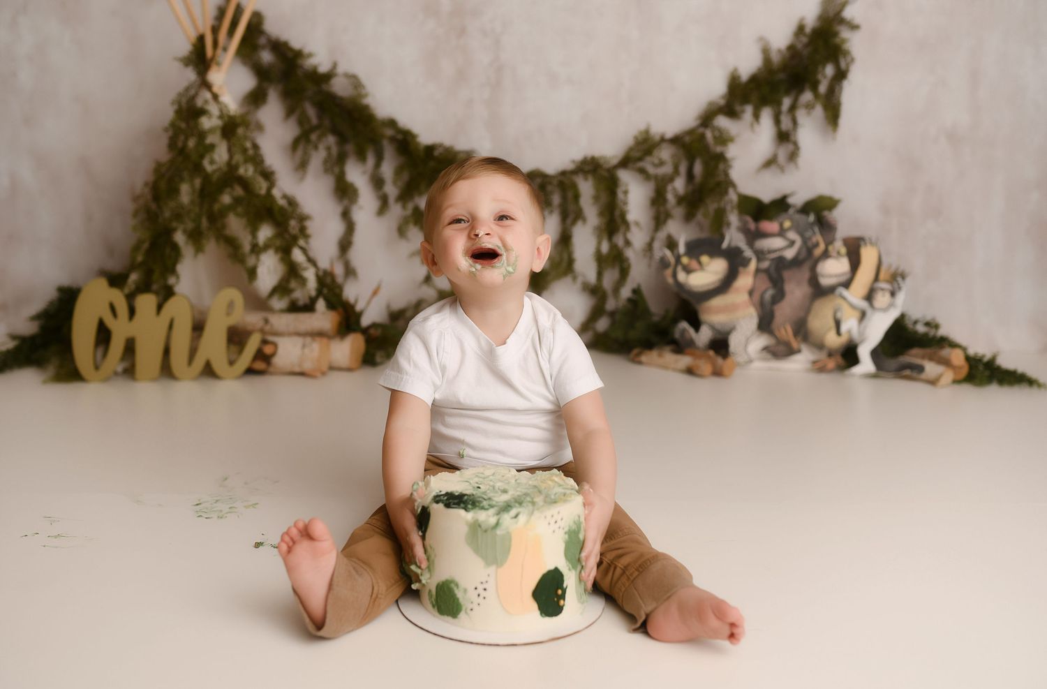 Laughing baby boy sits beside green leaf cake at woodland animal first birthday cake smash.