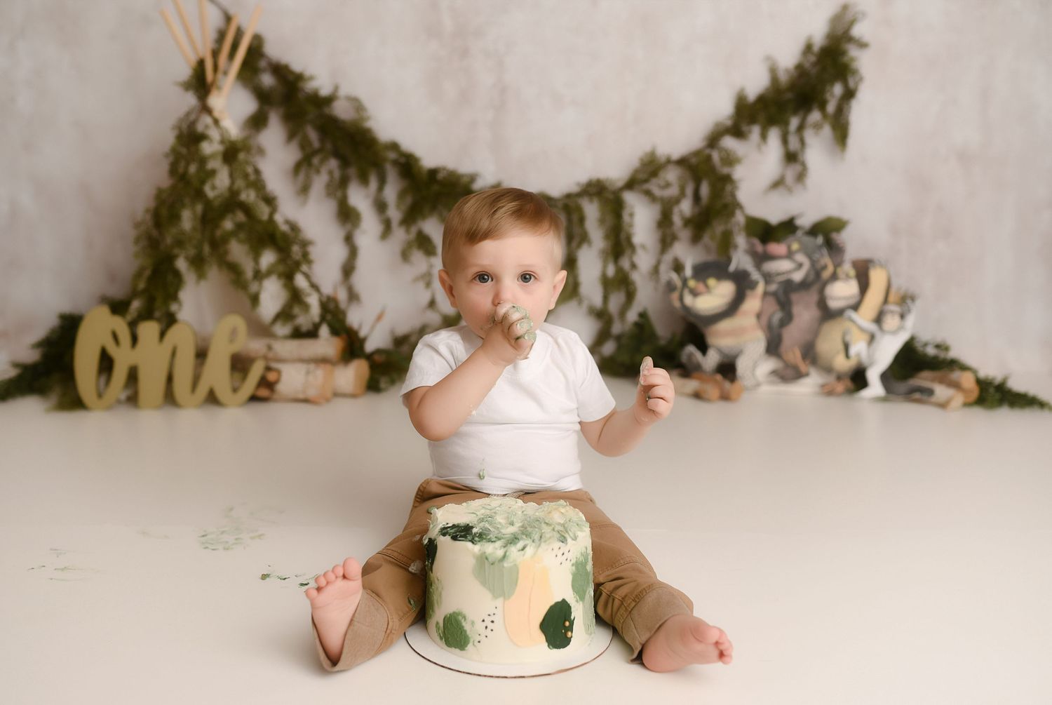 Baby boy in white onesie eats cake at woodland-themed first birthday cake smash photo session.